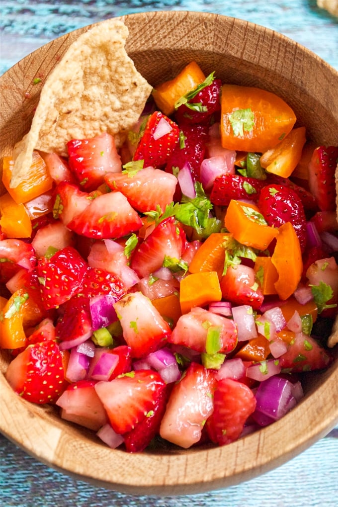 Close-up of strawberry salsa in a wooden bowl with a tortilla chip, featuring chopped strawberries, orange bell pepper, red onion, jalapeño, and cilantro.