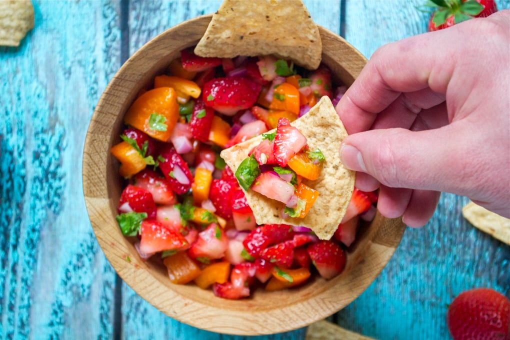 A hand holding a tortilla chip with strawberry salsa on it over a bowl of salsa.