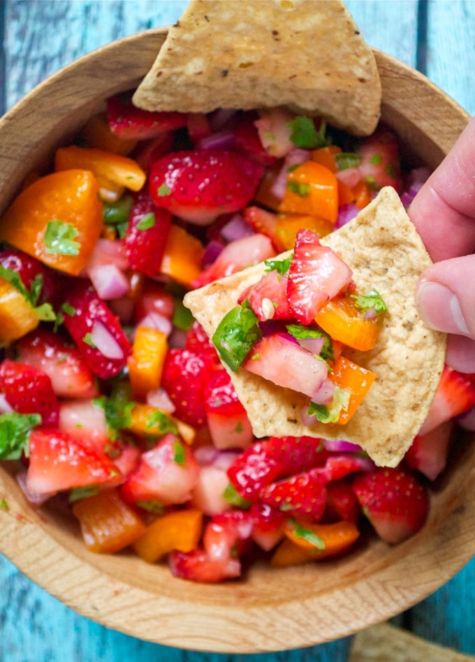 A hand holding a tortilla chip with strawberry jalapeño salsa over a  wood bowl.