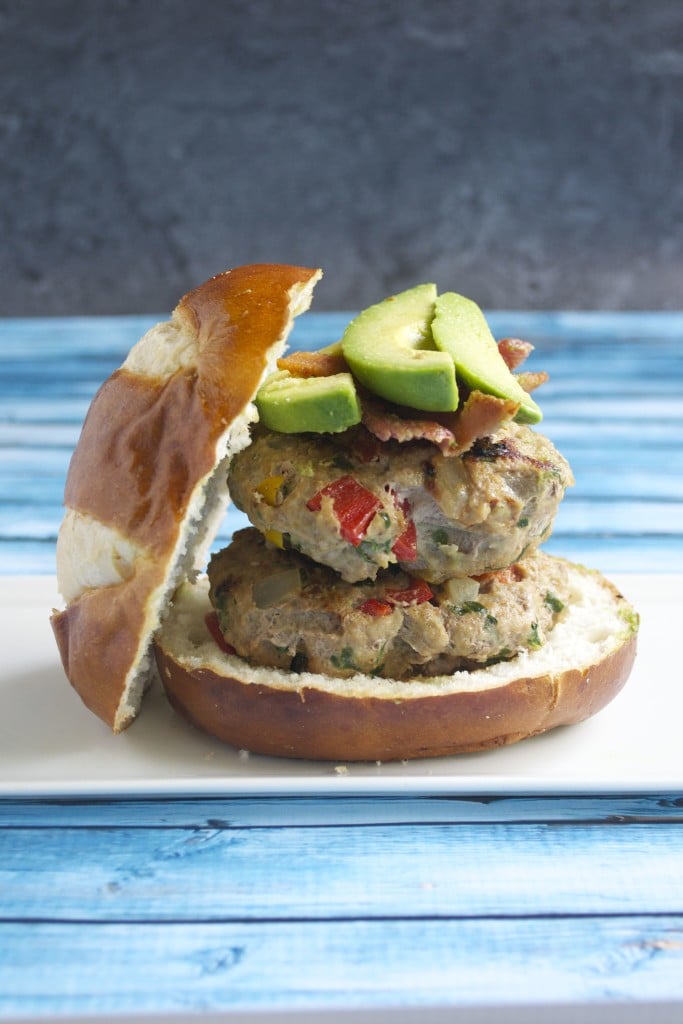 A stack of two veggie turkey burgers on a bun topped with sliced avocado and bacon. 