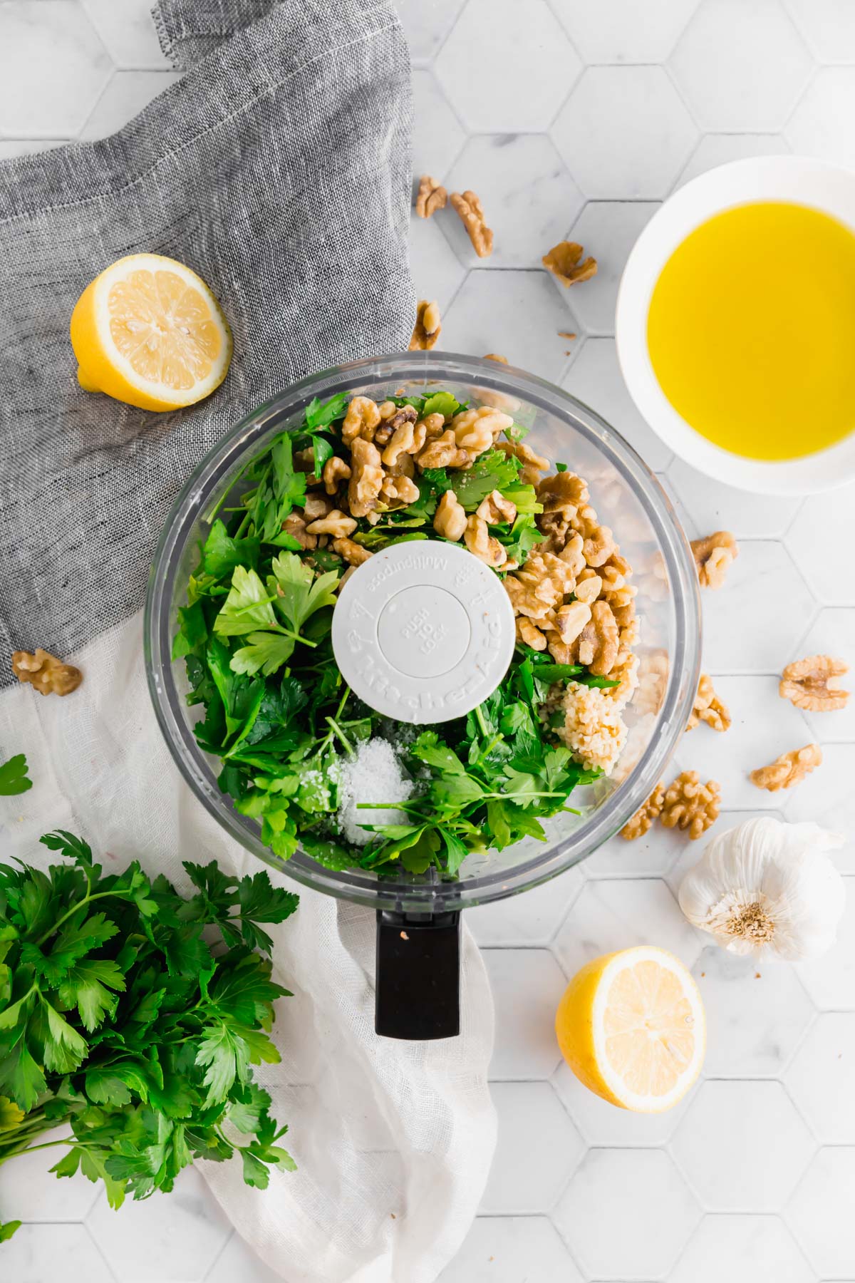 An overhead photo of a food processor filled with fresh parsley, garlic, walnuts, and salt to make vegan walnut parsley pesto.