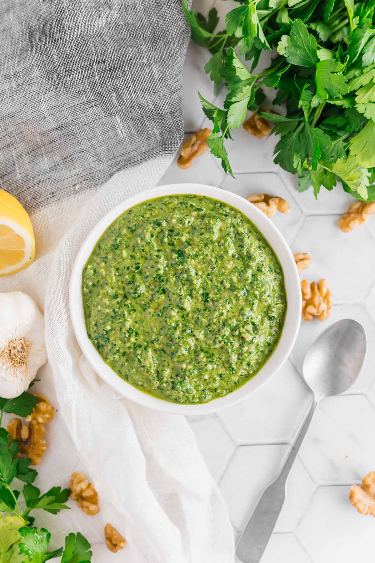 An overhead photo of a small bowl filled with vegan parsley walnut pesto with a spoon, fresh parsley, walnuts, garlic and lemon on a table.