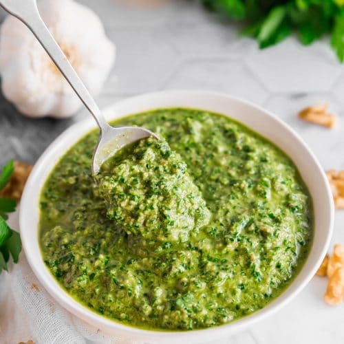 A photo of a bowl of vegan parsley walnut pesto with a spoon in it surrounded by a bulb of garlic, half a lemon, a bunch of parsley and walnuts on a table.