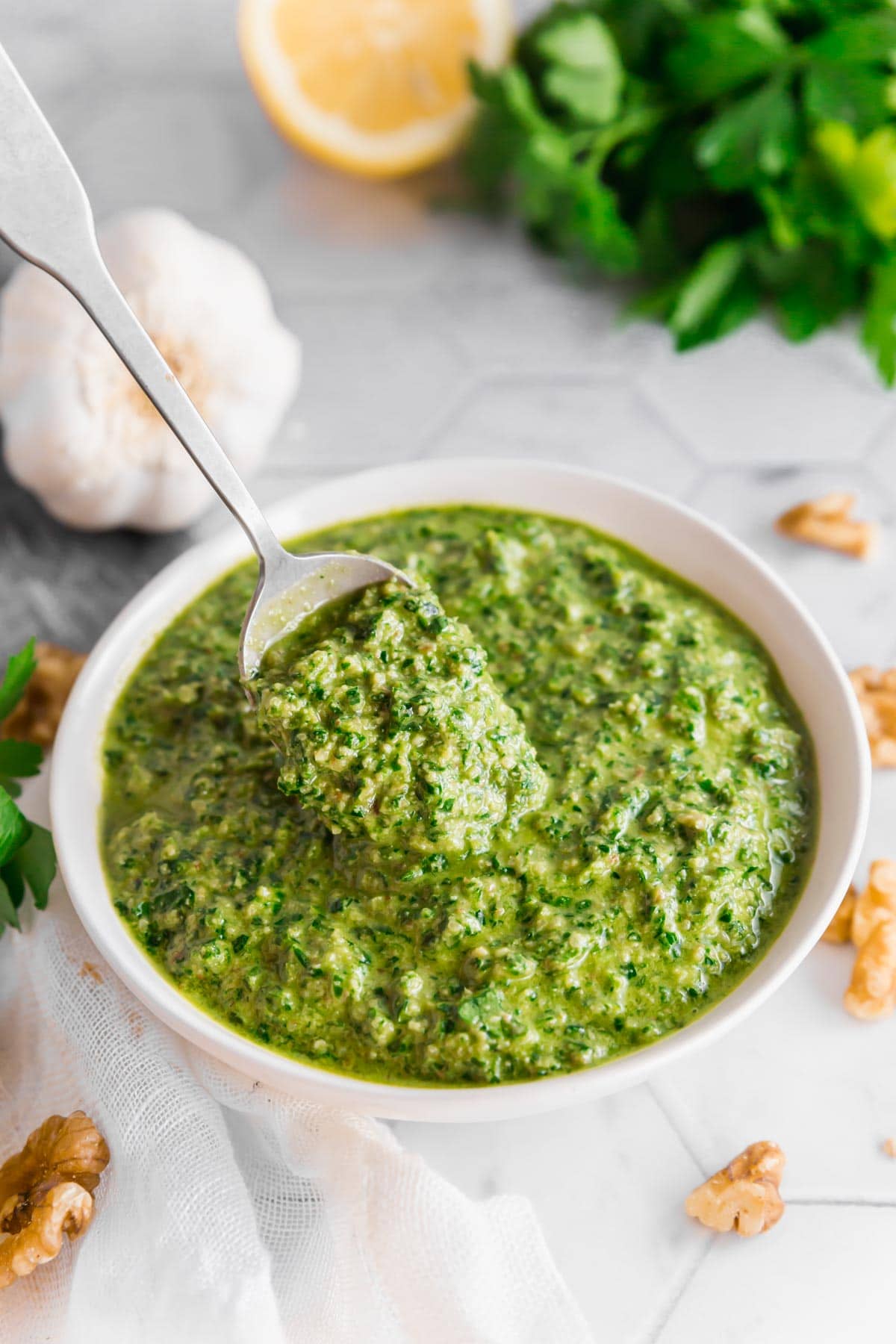 A photo of a bowl of vegan parsley walnut pesto with a spoon in it surrounded by a bulb of garlic, half a lemon, a bunch of parsley and walnuts on a table.
