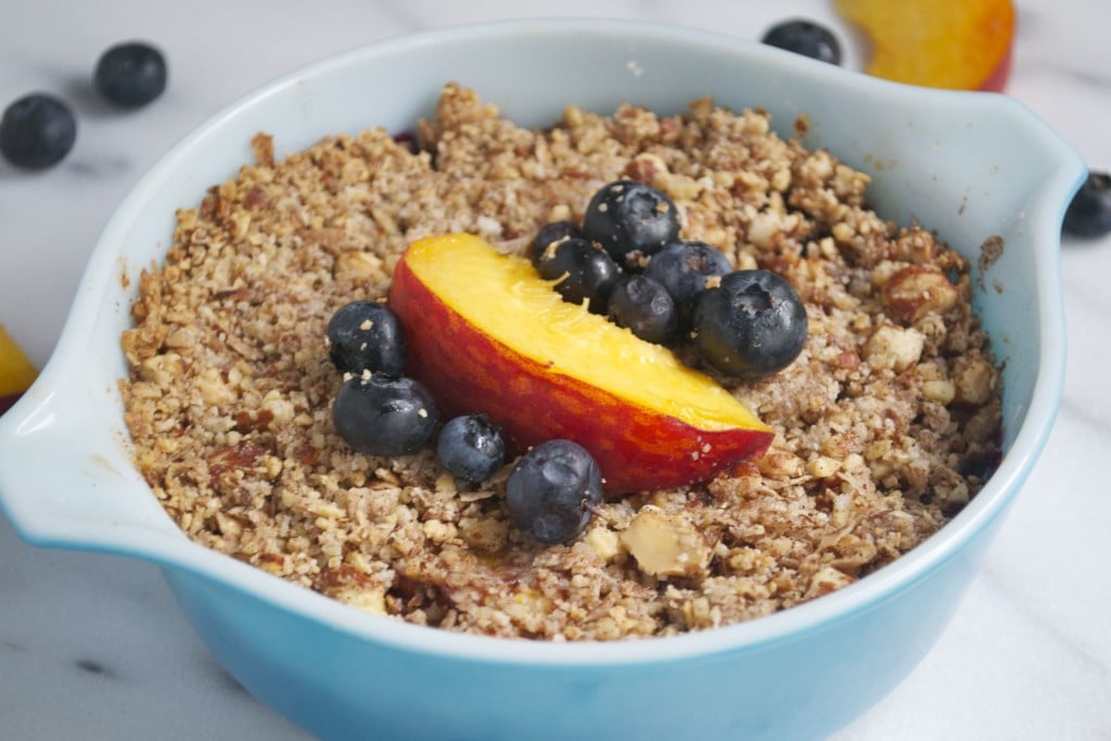 A ramekin of blueberry peach crisp with fresh peach wedge and blueberries on top.