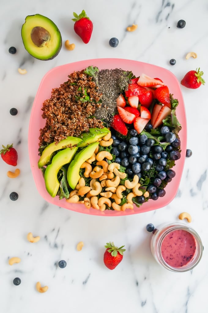 A pink serving bowl with quinoa, chia seeds, strawberries, blueberries, cashews, and avocado on a bed of lettuce.