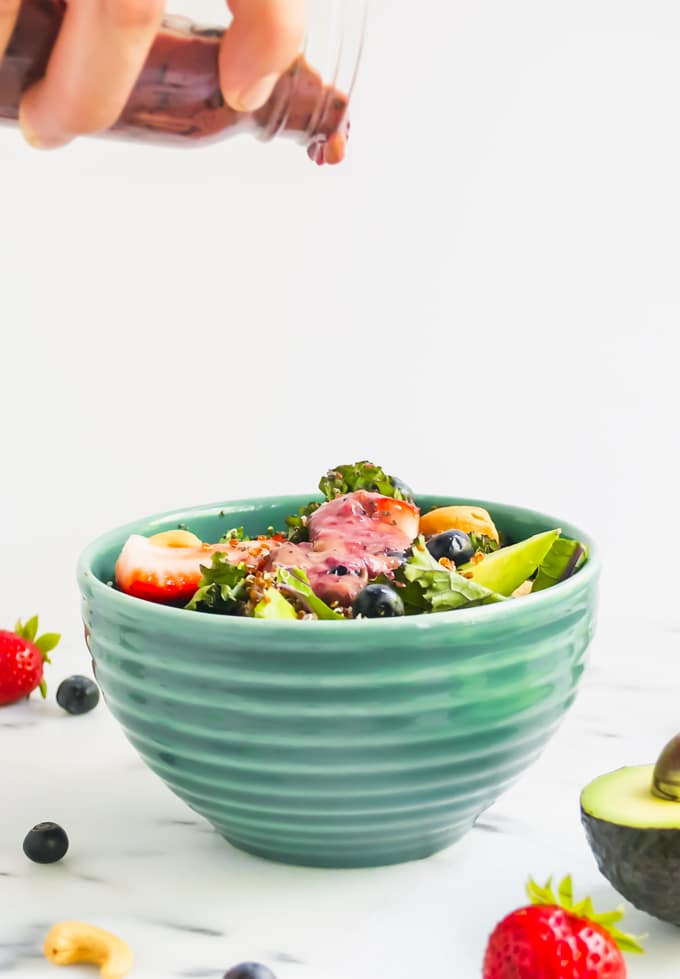 A hand pouring blueberry vinaigrette over a bowl of salad.