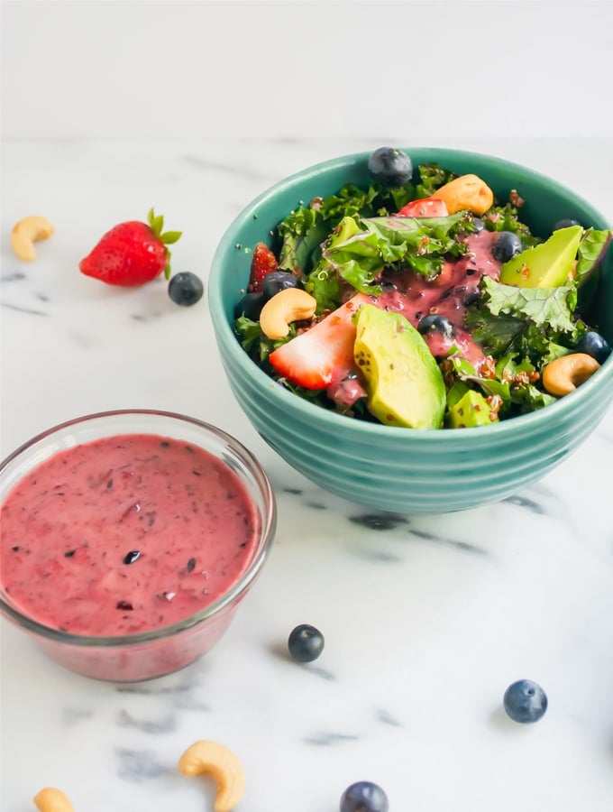 A bowl of salad with avocado, strawberries, kale, blueberries and cashews with a small bowl of blueberry vinaigrette on the side.