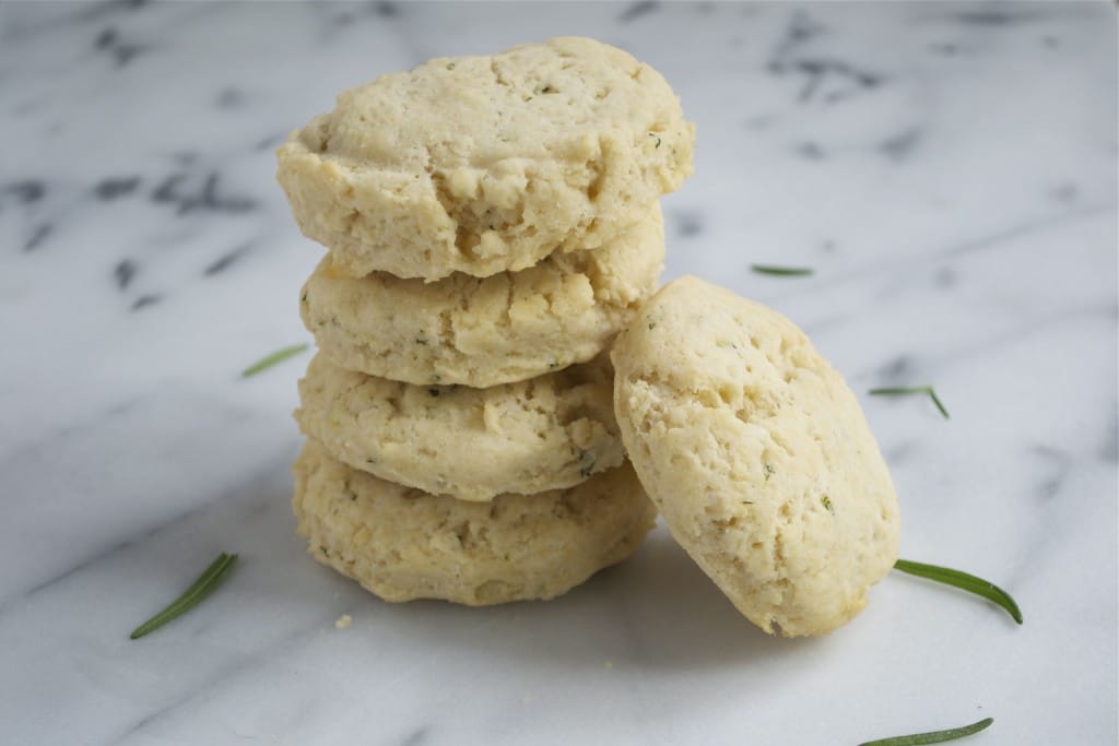 A stack of vegan rosemary biscuits with one leaning against the stack and fresh rosemary sprigs on the surface.