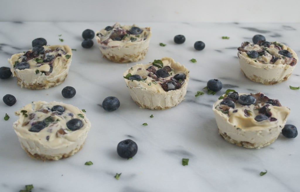 Mini vegan blueberry basil cheesecakes scattered on a marble table with fresh blueberries on the surface.