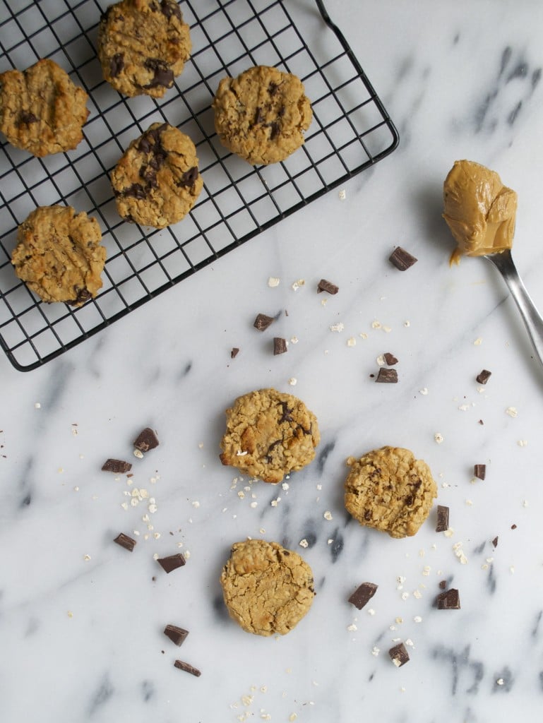 Vegan peanut butter chocolate chunk cookies on a cooling rack with additional cookies on a marble table scattered with chocolate chunks and a spoon of peanut butter.