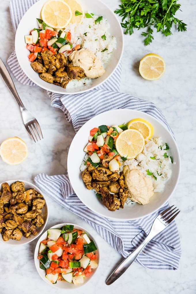 Mediterranean chicken bowls with spiced chicken thighs, creamy hummus, parsley lemon rice, and tomato cucumber parsley salad with fresh lime wedges. 