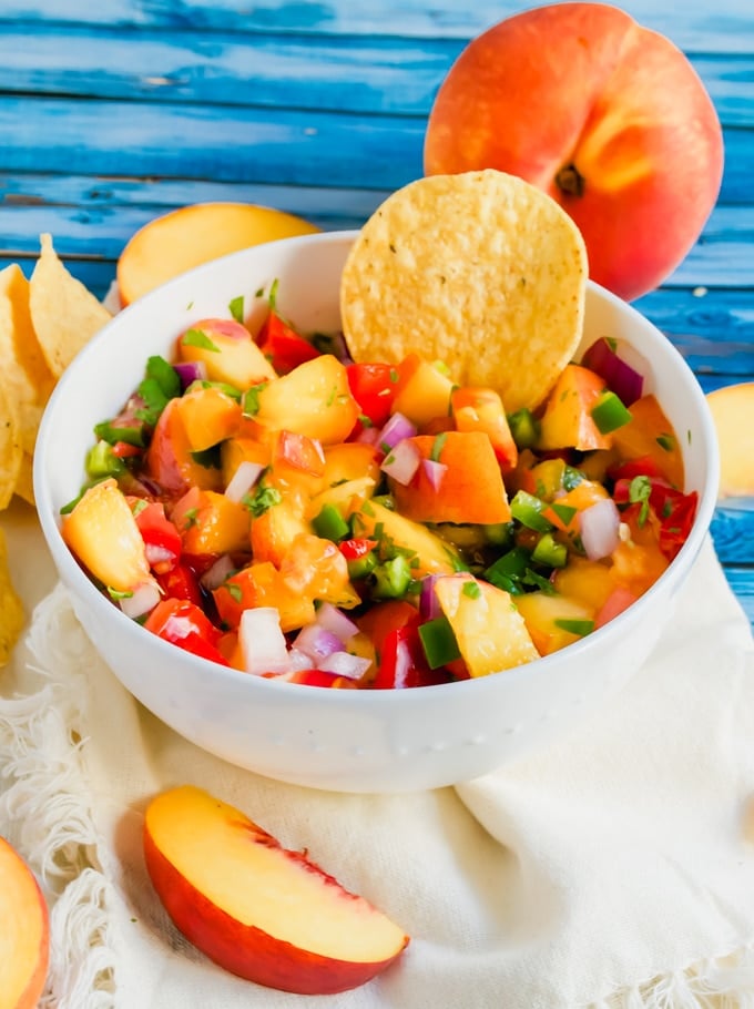 A bowl of peach tomato salsa with a tortilla chip in it and fresh peaches on the table.