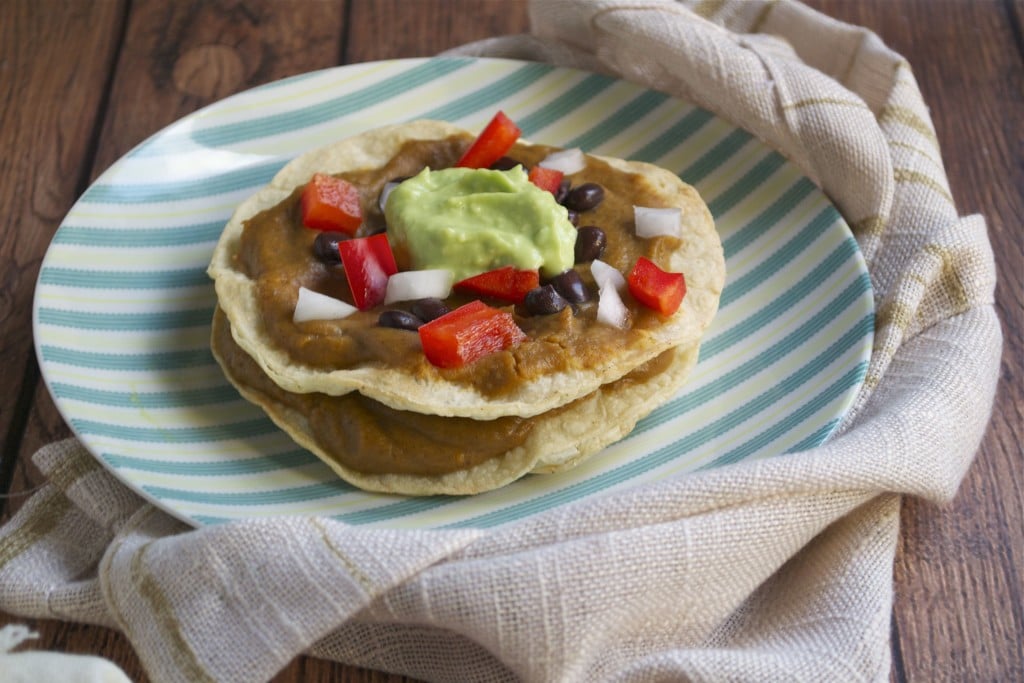 A stack of two sweet potato black bean tostadas on a plate topped with red bell pepper, red onion and avocado cream. 