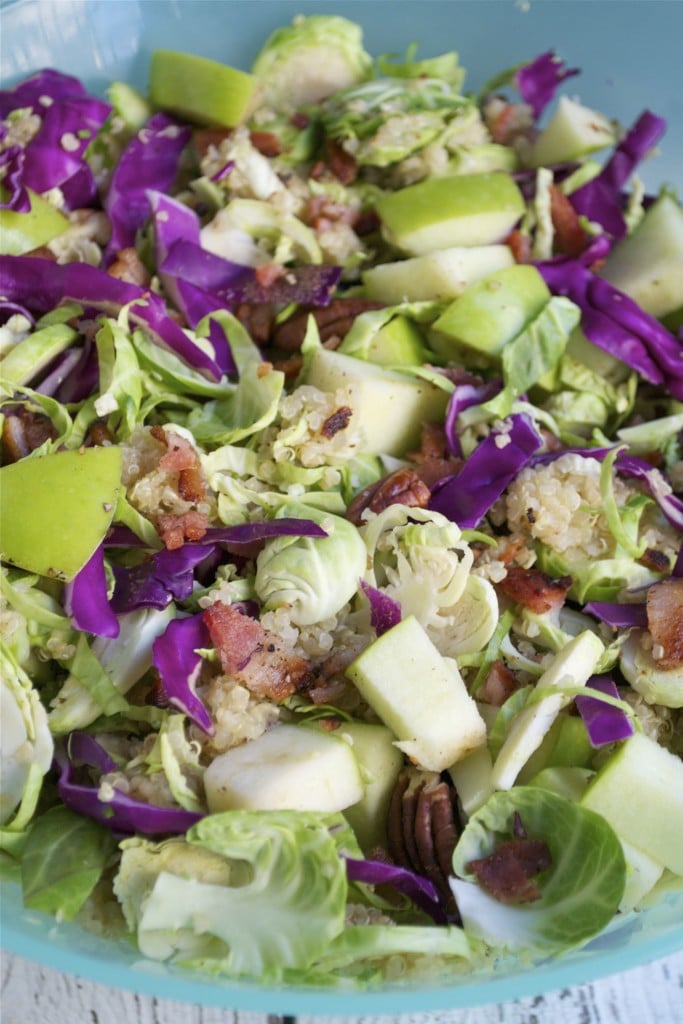 A closeup of shaved brussels sprouts salad topped with bacon bites, red cabbage, quinoa, pecans and diced apples.