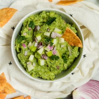 A bowl of green apple guacamole with baked tortilla chips, red onion, and limes.