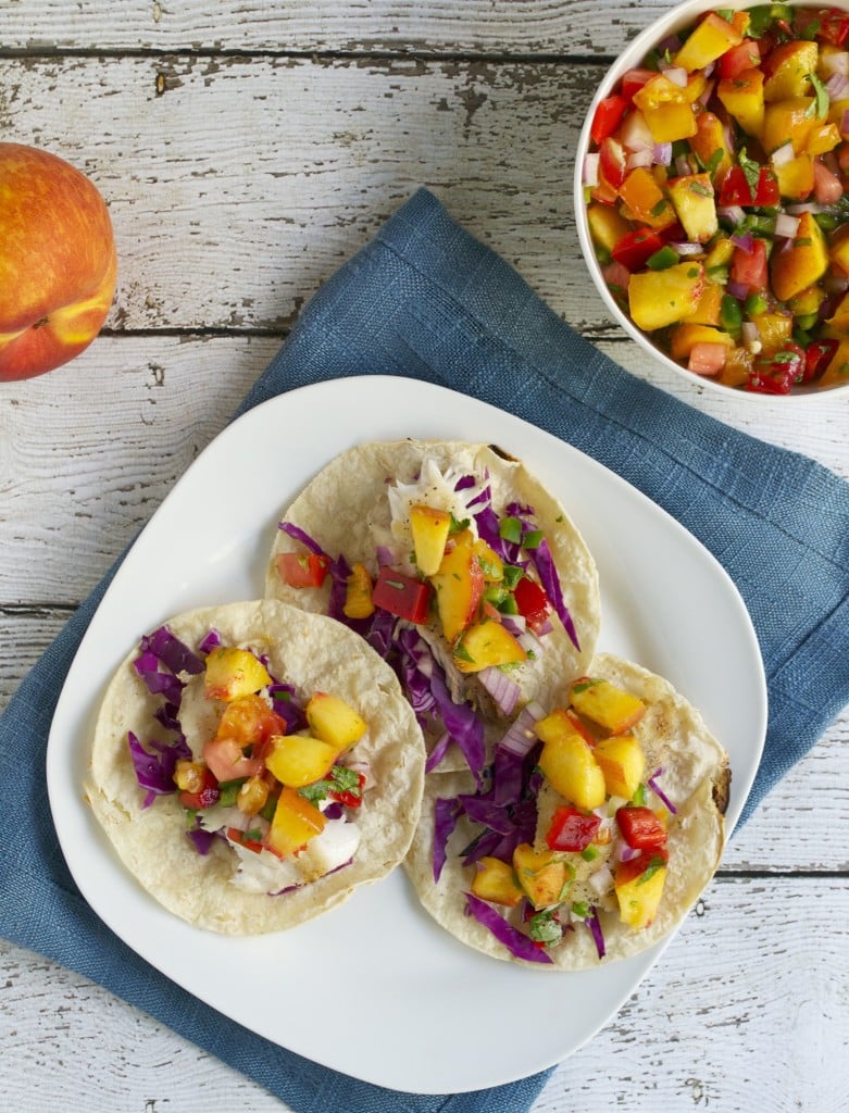 A plate of tilapia tacos topped with red cabbage, peach salsa, and tomatoes on a blue linen with a side of peach salsa.