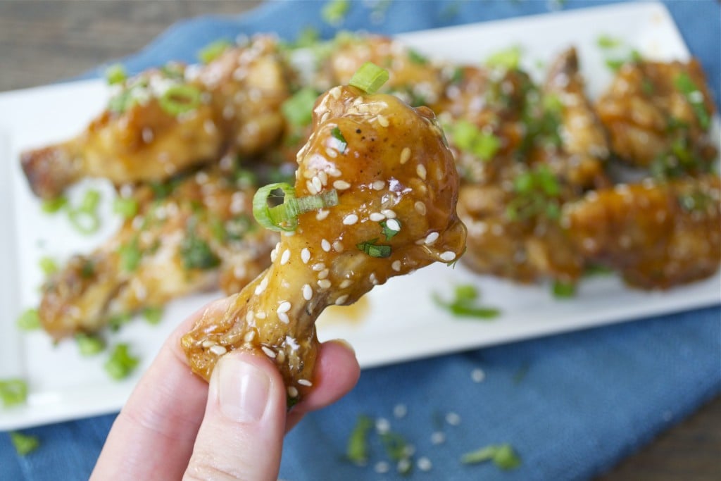 A hand holding a teriyaki chicken wing topped with green onion and sesame seeds.