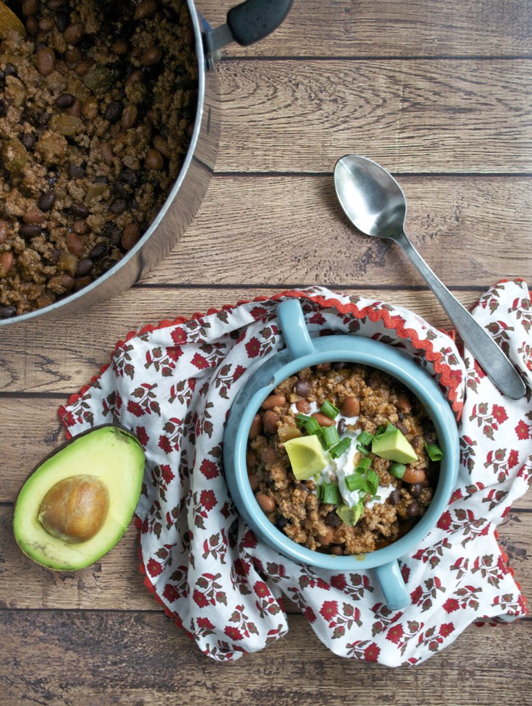An overhead view of a small crock of beef chili with half an avocado on the side and a large pot of chili next to it.