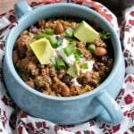 A blue bowl filled with Gluten-Free Beef Chili, topped with sour cream, diced avocado, and chopped green onions sits on a floral-patterned cloth with a halved avocado in the background.