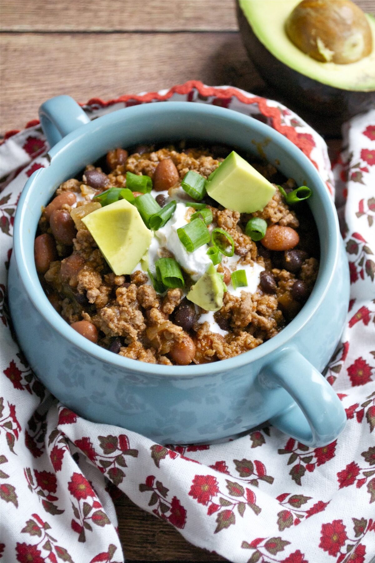 A blue bowl filled with Gluten-Free Beef Chili, topped with sour cream, diced avocado, and chopped green onions sits on a floral-patterned cloth with a halved avocado in the background.
