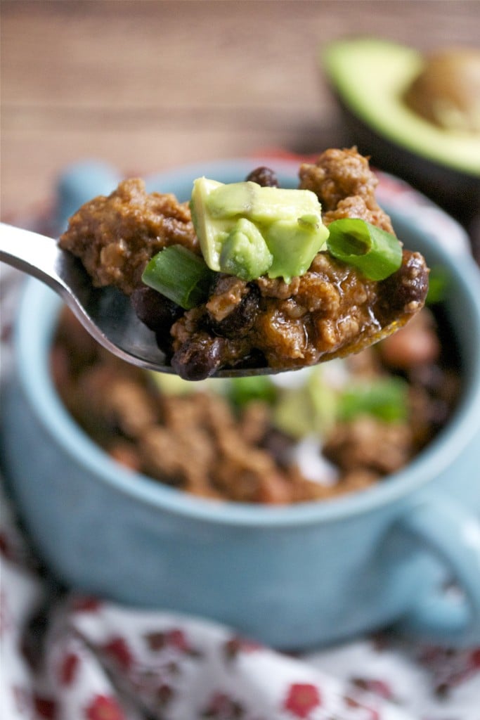 A spoon full of beef chili topped with green onions and diced avocado. 