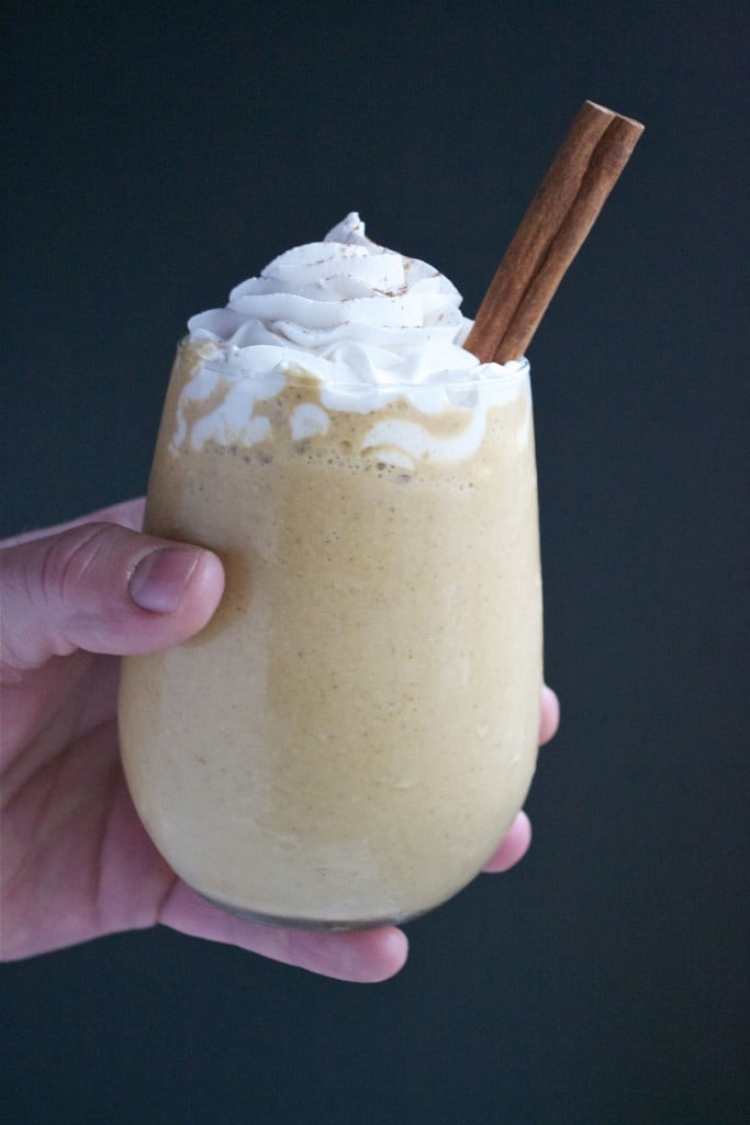 A hand holding a glass of pumpkin spice latte milkshake topped with a cinnamon stick and homemade whipped cream.