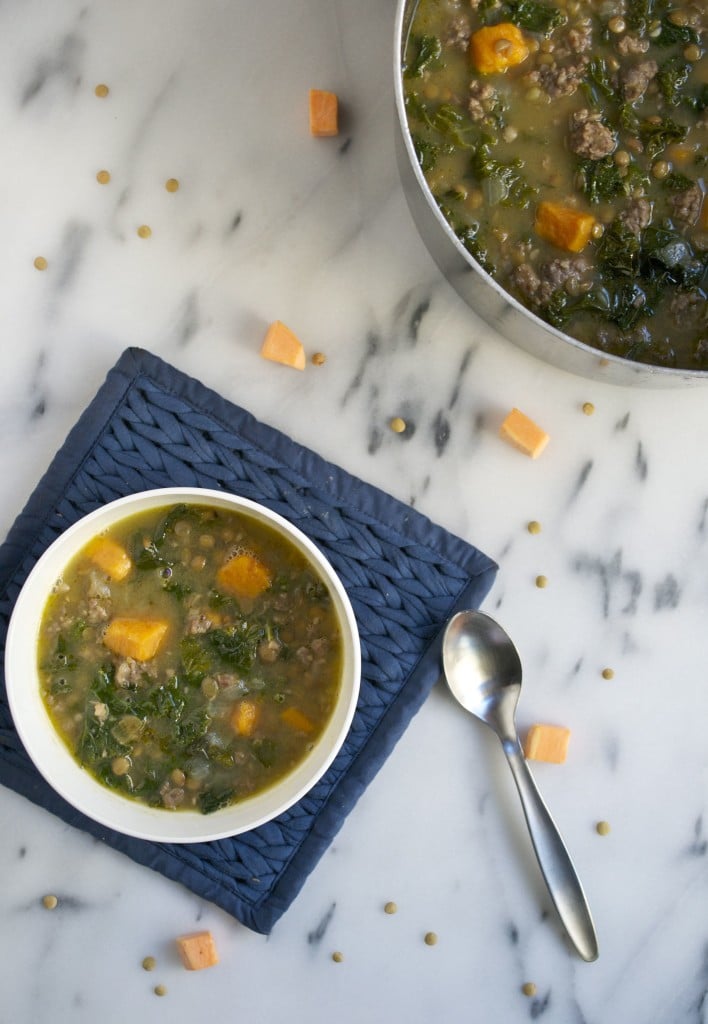 A bowl of kale, sausage, and lentil soup on a blue hot pad with a pot of soup on the side.