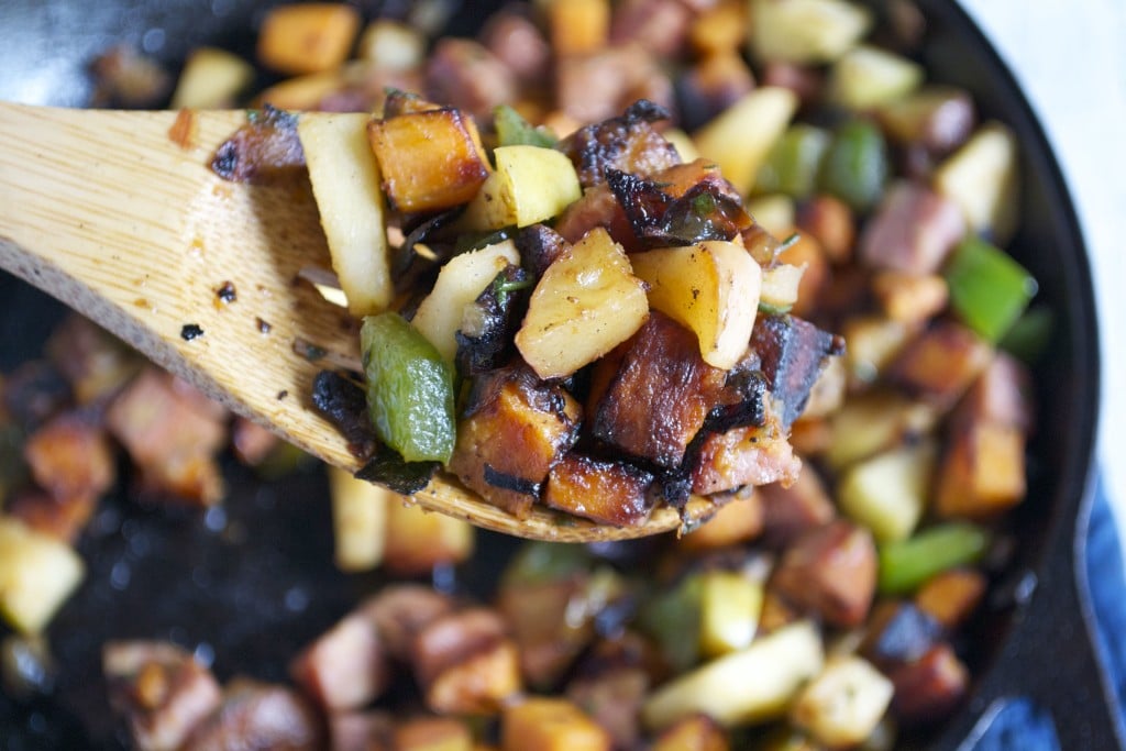 A wooden spoon with apple, sausage, sweet potato hash on it with a cast iron skillet beneath it.