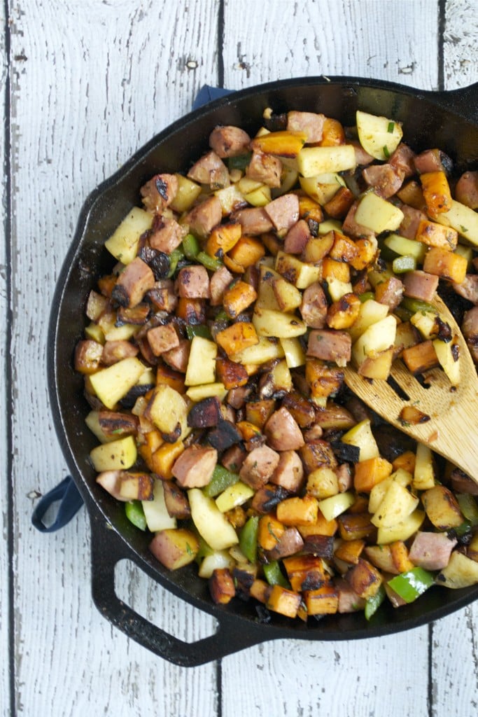 An overhead view of a cast iron skillet with apple, sausage, and sweet potato hash in it with a wooden spoon.