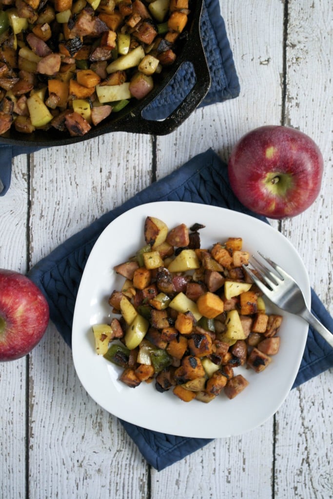 SausA white plate with apple, sausage, sweet potato hash with a fork and a cast iron skillet to the side.