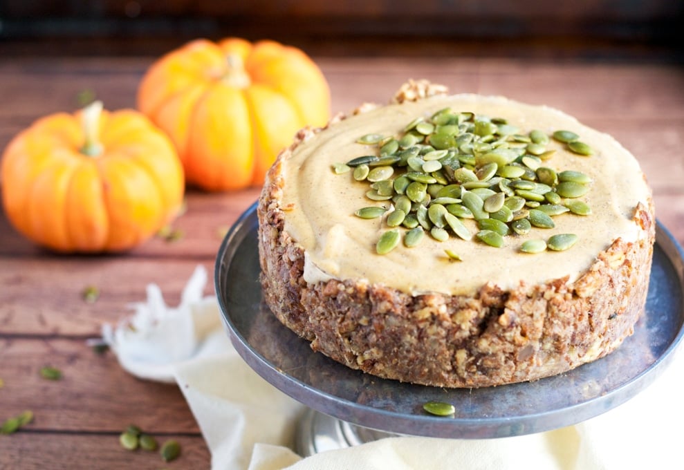 A small vegan pumpkin cheesecake topped with pumpkin seeds on a cake stand in front of two mini pumpkins.