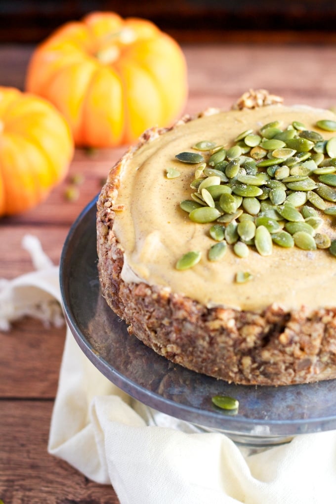 A small vegan pumpkin cheesecake with nut crust and topped with pumpkin seeds on a cake stand with mini pumpkins in the background.