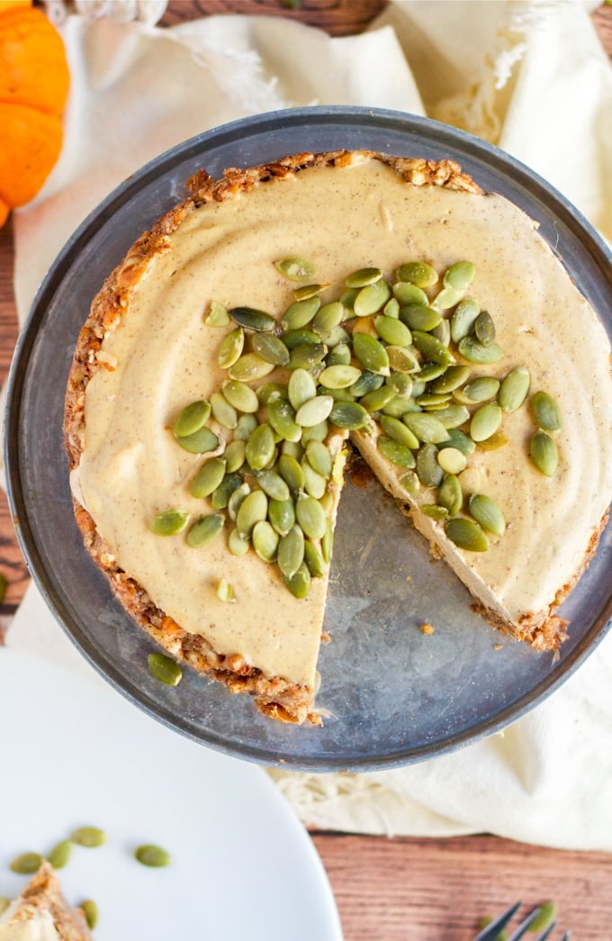 An overhead view of a vegan pumpkin cheesecake topped with pumpkin seeds with one slice removed.