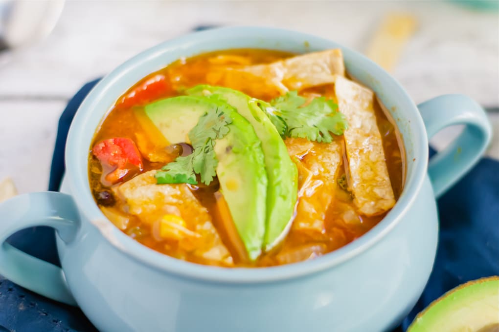 A blue soup bowl of chicken tortilla soup topped with tortilla strips, avocado slices and cilantro.