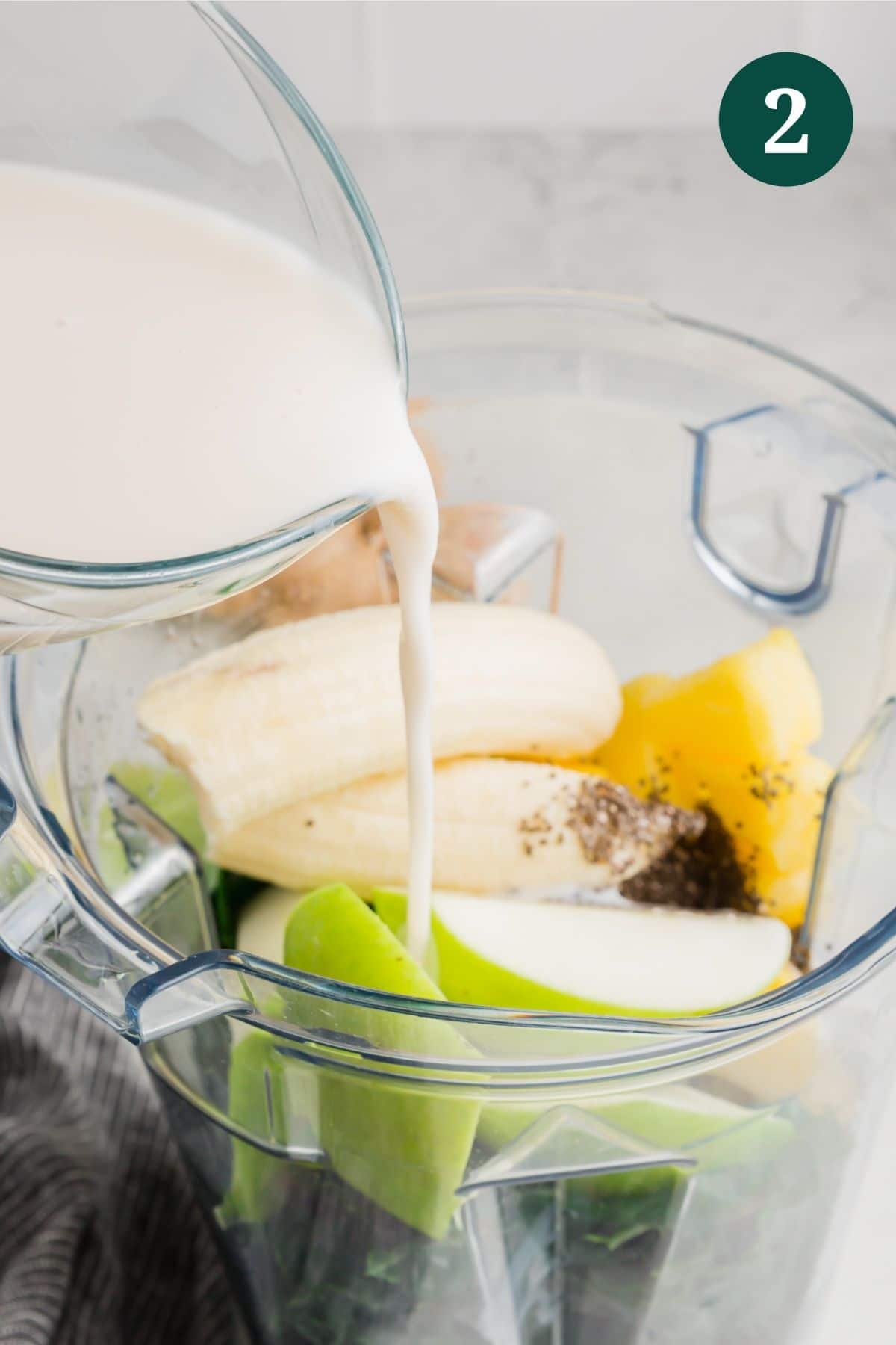 Almond milk being poured into a blend with apple, banana, pineapple and kale. 