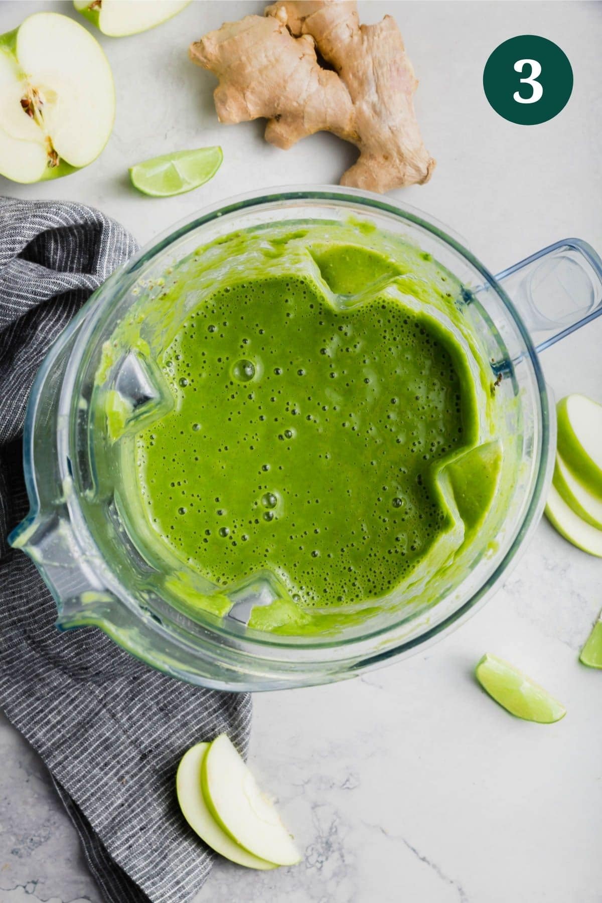 A blender with apple kale green smoothie in it and fresh ginger and apple on the table.