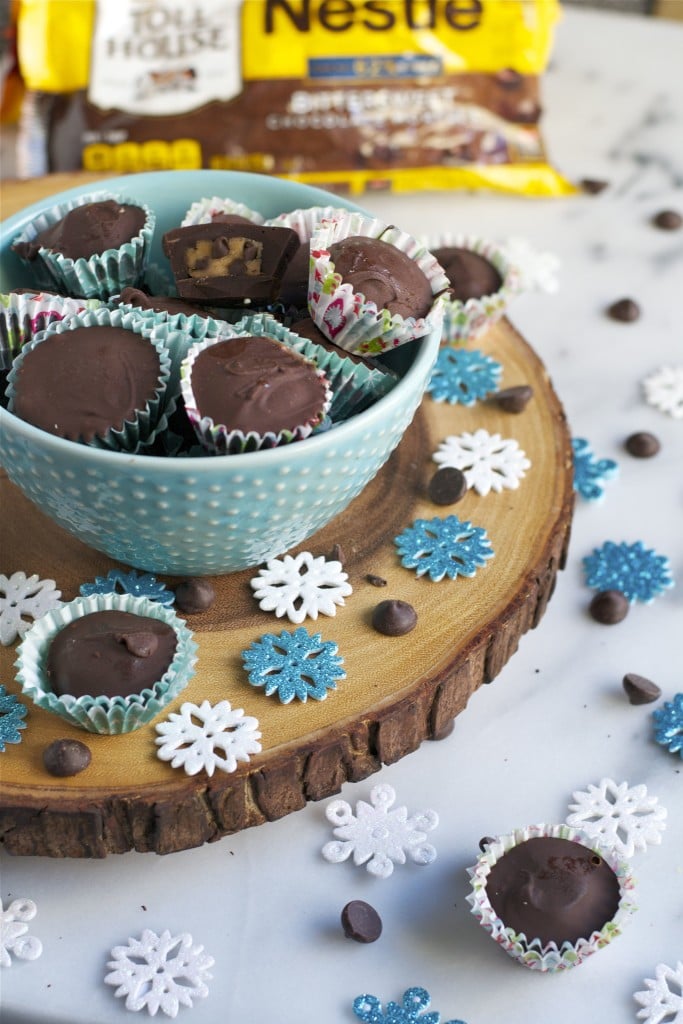 A bowl of chocolate chip cookie dough cups with paper snowflakes around the surface and a bag of chocolate chips in the background.