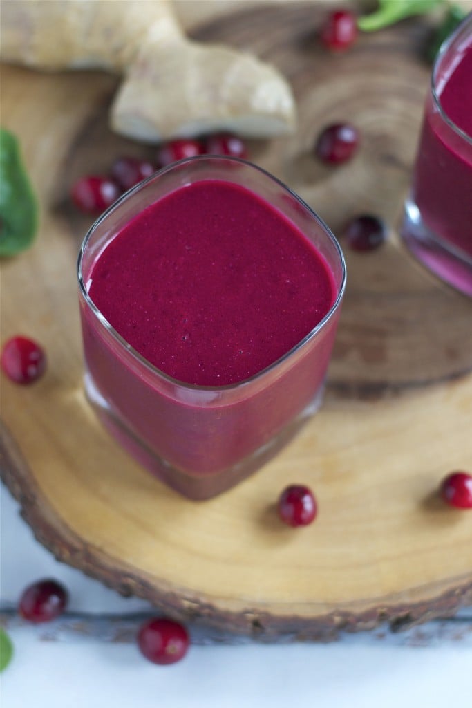 Glass of deep red cranberry beet smoothie on a wooden board, surrounded by scattered cranberries and fresh ginger in the background.