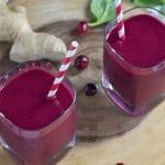 Overhead view of two glasses of cranberry beet smoothie with striped straws, surrounded by cranberries, spinach leaves, and fresh ginger on a round wooden board.