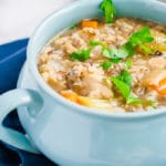 A light blue bowl filled with hearty Crockpot Chicken and Rice Soup, topped with fresh parsley, sits on a dark blue napkin with a gold spoon in the background.