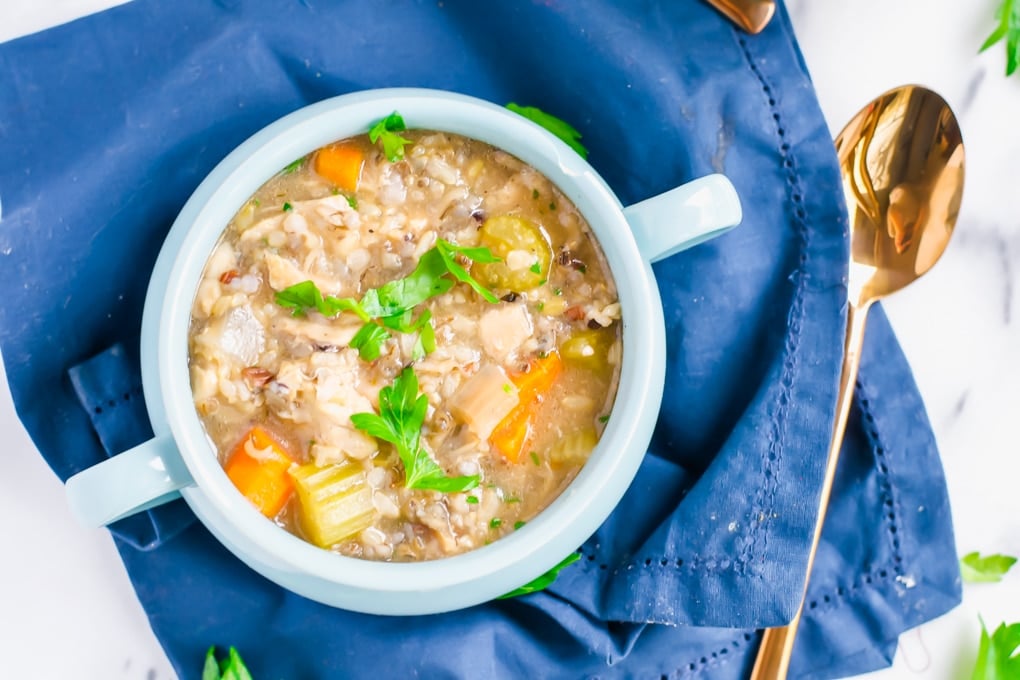 An overhead view of a bowl of chicken and rice soup with carrots, celery and fresh parsley with a spoon on the side.