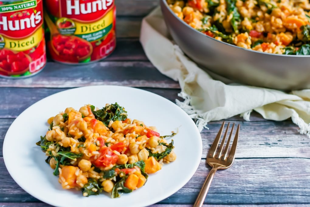 A plate of chickpea kale sweet potato rice skillet with a skillet and cans of diced tomatoes in the background.