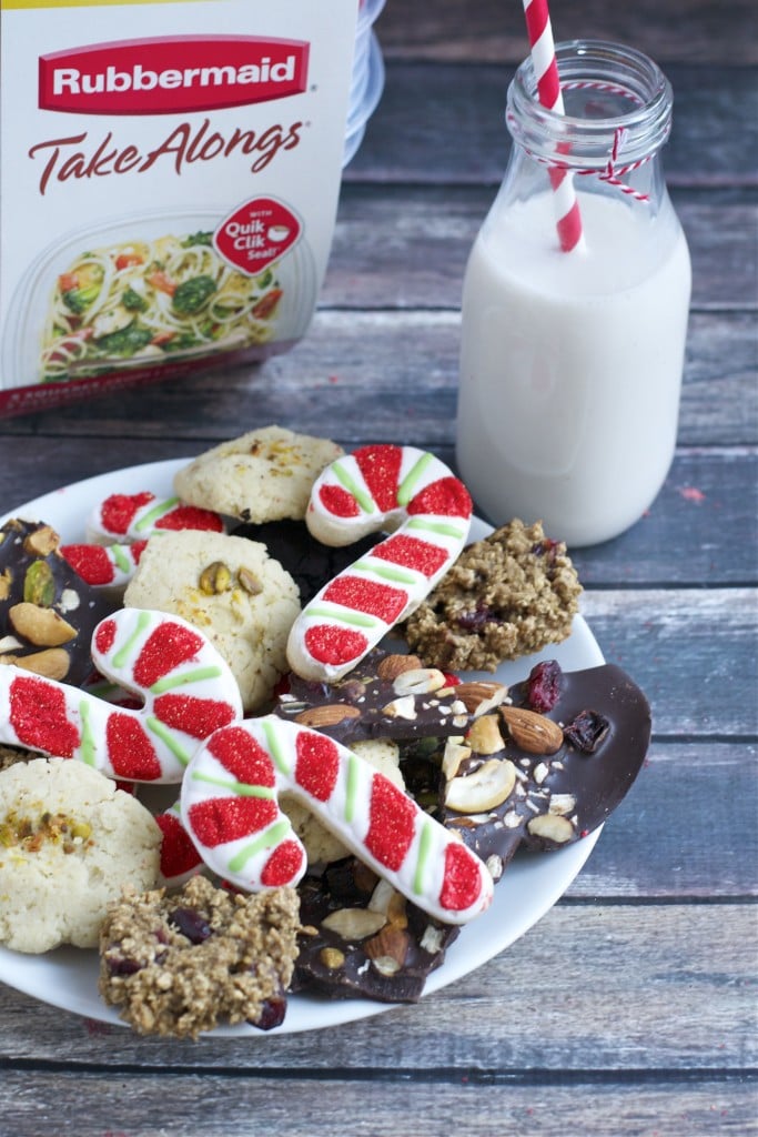 A plate of assorted cookies with a glass of milk and tupperware containers in the background.