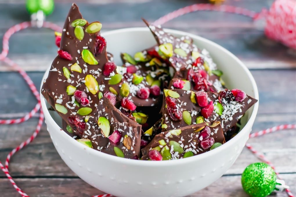 A bowl of pomegranate pistachio chocolate bark with mini ornaments and red twine around it.