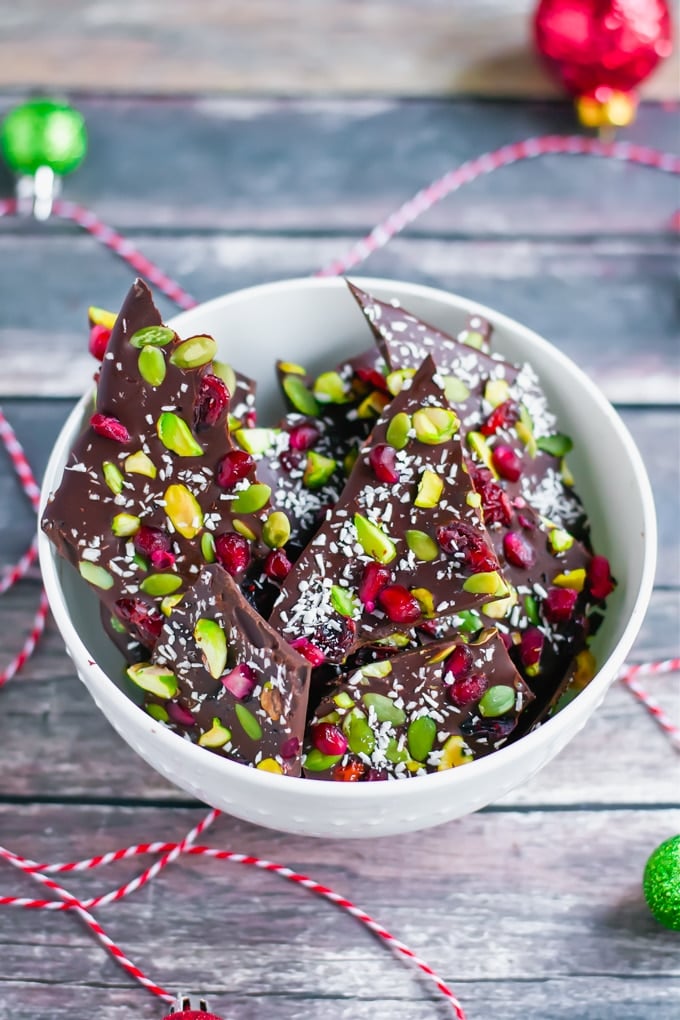A bowl of pistachio and pomegranate chocolate bark with red twine and mini ornaments around it.