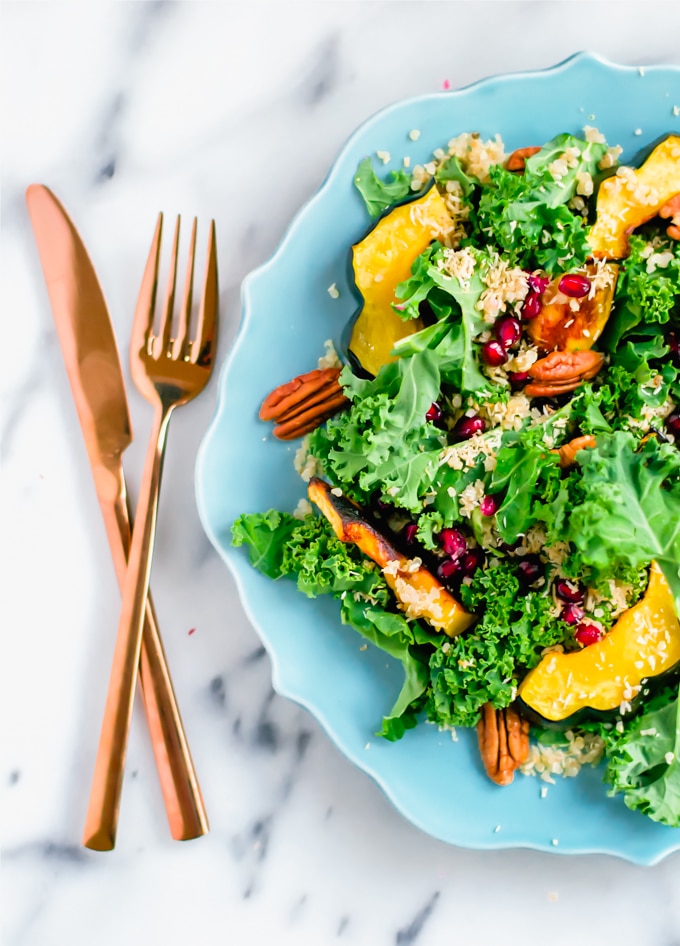 A blue plate with kale salad topped with roasted acorn squash, pecans, pomegranate arils, and toasted coconut with a fork and knife to the side.