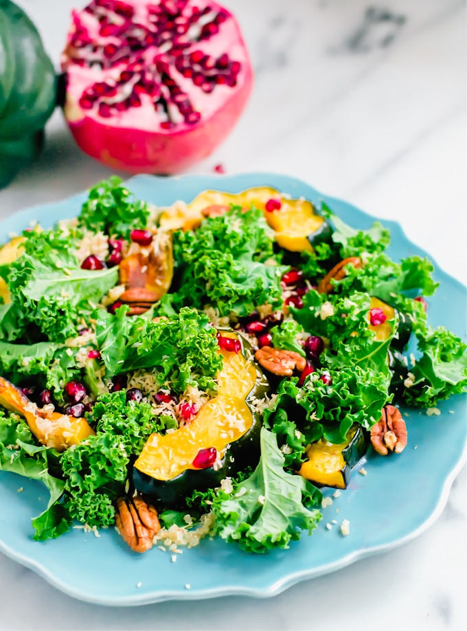 A plate of kale and acorn squash salad topped with pecans and pomegranate arils with fresh pomegranate in the background.