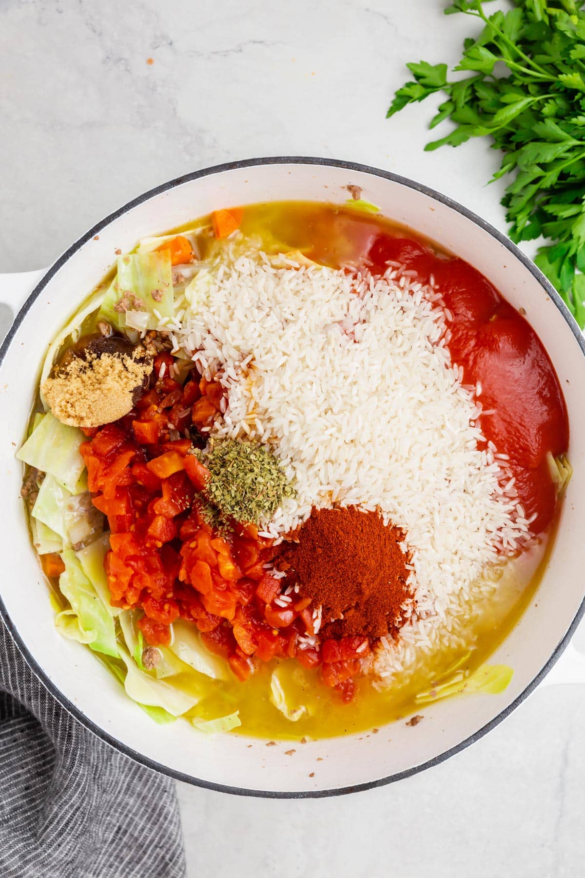 A white dutch oven with chicken broth, uncooked rice, tomato paste, dried oregano, dried paprika, brown sugar, diced tomatoes, and shredded cabbage before mixing together.