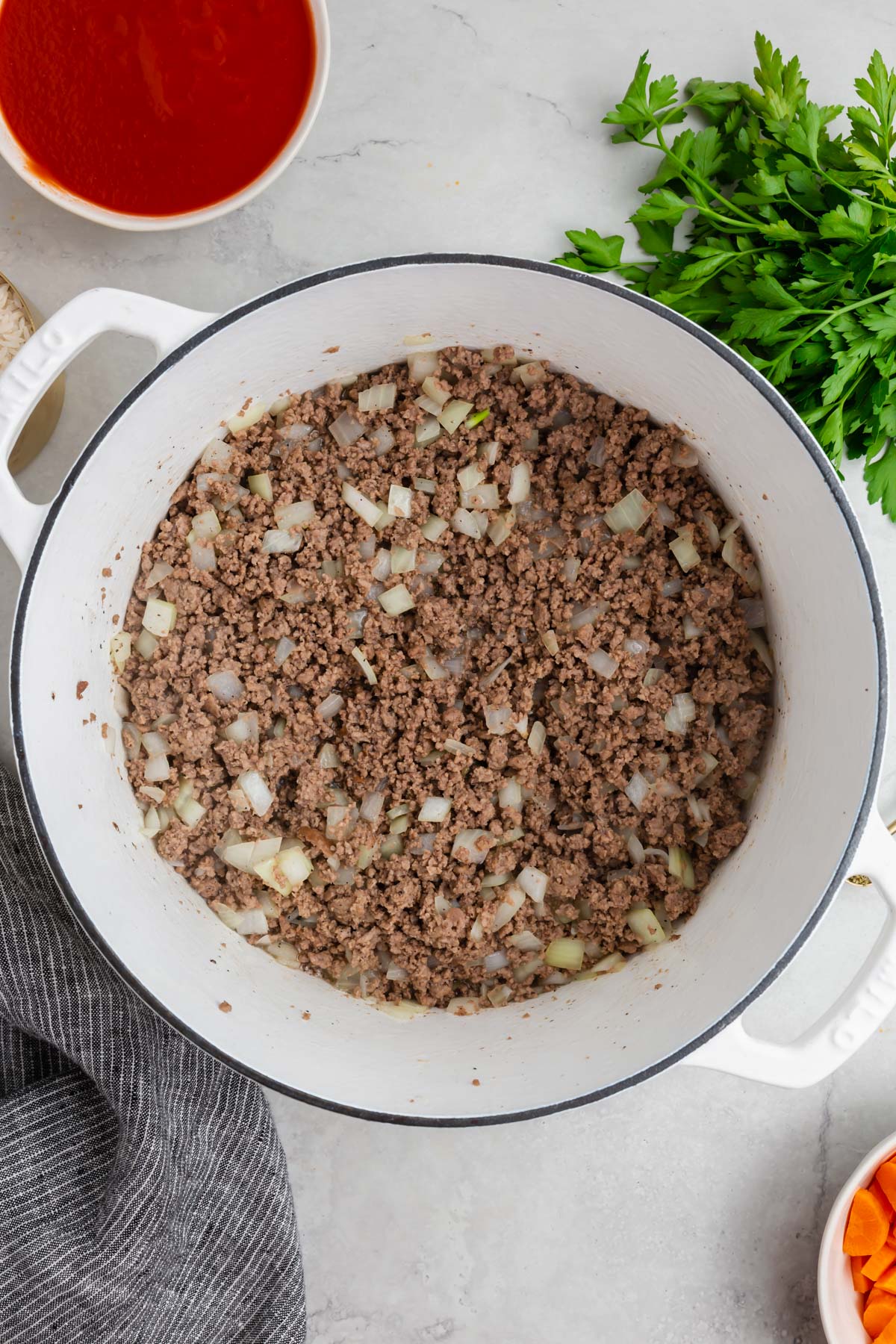 A white dutch oven with cooked ground beef mixed with diced onions and a bowl of tomato paste to the side.